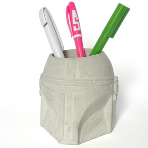 May include: A grey concrete pencil holder shaped like a Star Wars Mandalorian helmet. The holder has three pens inside: a pink pen, a green pen, and a white pen.