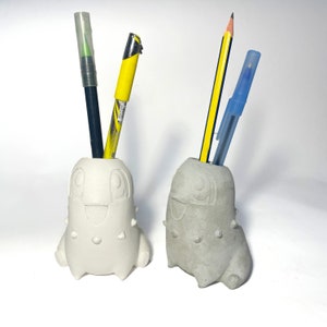 Puede incluir: Dos portalápices con forma de Chikorita, uno blanco y otro gris, cada uno con bolígrafos y lápices. El soporte blanco contiene un bolígrafo negro y otro amarillo. El soporte gris contiene un lápiz y un bolígrafo azul. Sobre una superficie blanca.