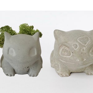Puede incluir: Dos macetas de hormigón gris con forma de personaje Pokemon, una con musgo verde en su interior.