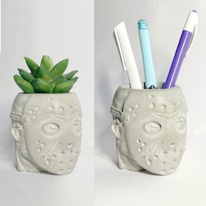May include: A gray concrete planter shaped like a hockey mask with a green succulent plant inside. The planter is also used as a pencil holder with three pens inside.