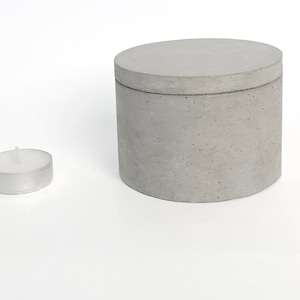 Puede incluir: Un recipiente cilíndrico de hormigón con tapa a juego, junto a una pequeña vela de té. El recipiente es de color gris claro y tiene una superficie texturizada. La vela está en una taza de metal plateado.