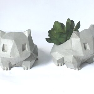 May include: Two grey concrete planters shaped like a cartoon animal, one with a green succulent plant inside.
