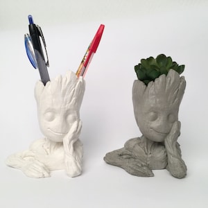 Puede incluir: Dos macetas de Groot de hormigón, una con un bolígrafo rojo y azul y la otra con una pequeña planta suculenta verde.