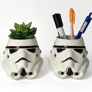 Può includere: Due organizer da scrivania bianchi a forma di casco da Stormtrooper. Uno contiene una succulenta verde, l'altro penne e un pennarello. I caschi hanno dettagli neri su sfondo bianco.