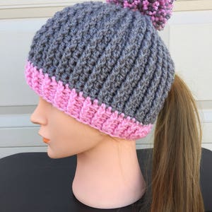 May include: Grey and pink crocheted beanie with a ponytail hole and a fluffy pom pom on top.