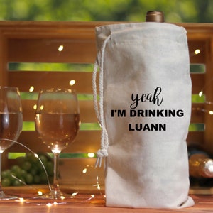Può includere: Una borsa per vino in tela bianca con un cordoncino nero. La borsa ha la scritta "yeah I'm drinking Luann" stampata su di essa.
