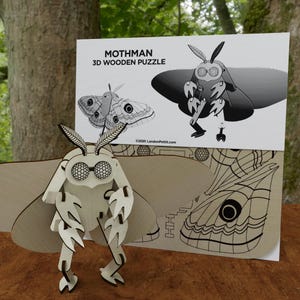 Puede incluir: Un rompecabezas 3D de madera de Mothman. El rompecabezas ensamblado es de color marrón claro con detalles en negro, con una criatura parecida a una polilla con ojos grandes y alas detalladas. El embalaje incluye ilustraciones y el texto "Mothman 3D Wooden Puzzle".