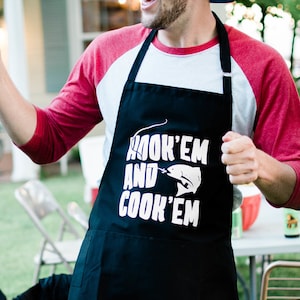 May include: A man wearing a red and white baseball shirt, a blue baseball cap, and a black apron with the text "Hook 'em and Cook 'em" and a fish graphic.
