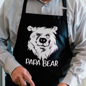 May include: Black apron with a white bear graphic and the text "Papa Bear". A person is wearing the apron and holding a knife.
