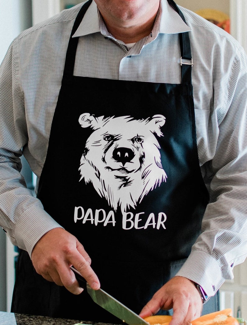 May include: Black apron with a white bear graphic and the text "Papa Bear". A person is wearing the apron and holding a knife.