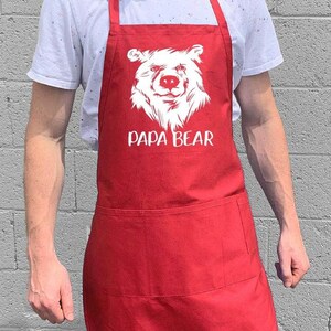 May include: Red apron with a white bear face and the text "Papa Bear" printed on the front. The apron has a large pocket on the front.