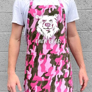 May include: Pink and green camouflage apron with a white bear graphic and the text "Papa Bear".