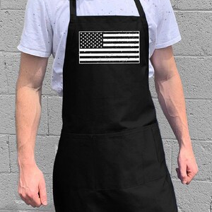 May include: A black apron with a white distressed American flag design. The apron has a large pocket on the front.