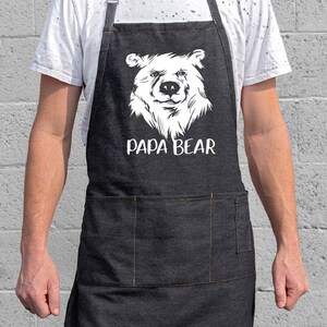May include: A black denim apron with a white bear face and the text "Papa Bear" printed on it. The apron has a large pocket on the front.