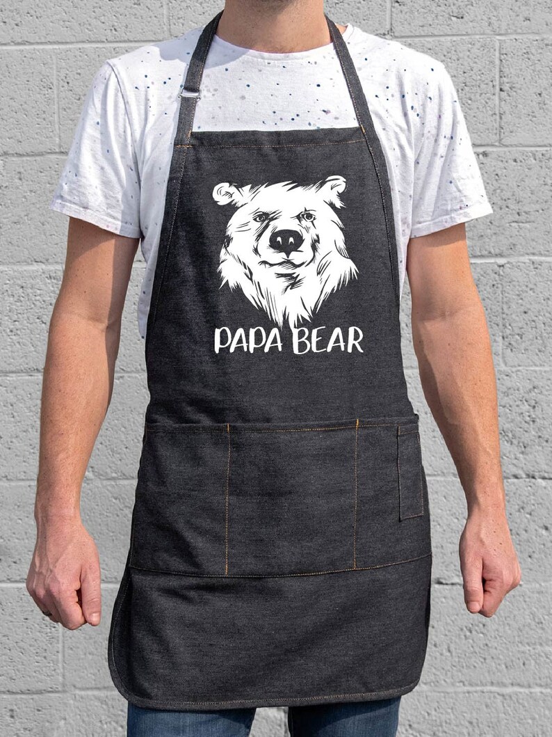 May include: A black denim apron with a white bear face and the text "Papa Bear" printed on it. The apron has a large pocket on the front.