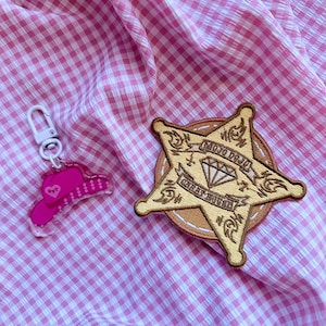 May include: A pink and white gingham fabric background with a pink cowboy hat charm and a beige embroidered sheriff's star patch. The star patch has the text "MOJO DOLL" and "COWGIRL HONKY" with a diamond in the center.