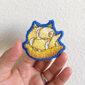 May include: Embroidered patch featuring a yellow and white sailboat with gold accents, set against a blue background. The patch has a detailed design and is likely intended for clothing or accessories.