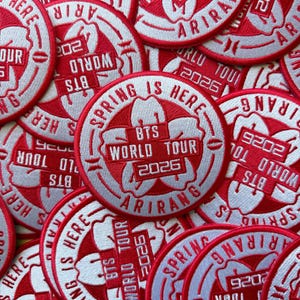 May include: A collection of red and white embroidered patches. The patches feature the text "BTS WORLD TOUR 2026 ARIRANG" and "SPRING IS HERE" in a circular design. The patches are likely for a concert or event.