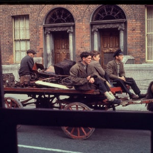 May include: Four men are sitting on a horse-drawn cart filled with scrap metal. The cart is on a paved street in front of a brick building with two doorways. The men are wearing casual clothing and hats. The horse is brown and has a black harness.