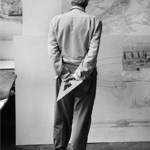 May include: A black and white photograph of a person standing with their back to the camera, wearing a long-sleeved button-down shirt and trousers. The person holds a clear plastic triangular drafting ruler in their hands behind their back.