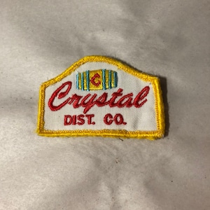 May include: A white embroidered patch with a yellow border. The patch features the word "Crystal" in red cursive lettering and the words "Dist. Co." in black lettering below. The patch also features a stylized image of a barrel with a blue and yellow design.