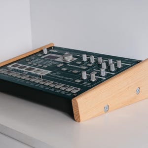 May include: Electronic music synthesizer with a dark teal control panel and silver knobs. The synthesizer is set in a light wood frame. The device is on a white surface.