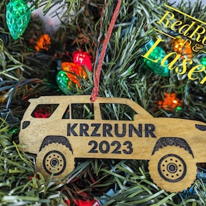 May include: Wooden Christmas ornament shaped like a car with the text "KRZRUNR 2023" on it. The ornament is hanging on a Christmas tree with red and green lights.