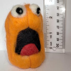 May include: Orange felted creature with wide eyes and an open mouth, approximately 2 inches tall. The mouth is black with a red interior. White felt is used for the eyes. A ruler is in the background.