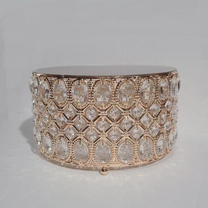 May include: A gold-colored, round cake stand with a mirrored top. The sides are adorned with clear, oval-shaped crystals set in a decorative pattern. The stand has a small, rounded foot.