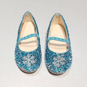 May include: A pair of light blue sparkly shoes with a silver snowflake design. The shoes have a strap across the top and are made for a child.