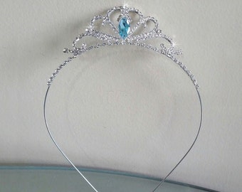May include: Silver tiara with a light blue jewel in the center and rhinestones around the crown. The tiara is on a silver headband.
