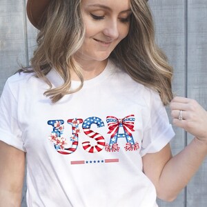 May include: A white t-shirt with a red, white, and blue USA graphic featuring flowers and bows. The graphic includes the word "USA" with stars and stripes.