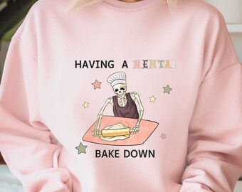 May include: Pink sweatshirt with a skeleton baking a cake and the text "Having a mental bake down".