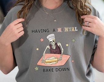 May include: Gray t-shirt with a skeleton baking a cake. The text on the shirt reads "Having a mental bake down."