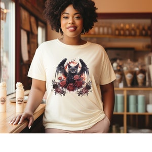May include: Cream-colored t-shirt featuring a black cat with bat wings, surrounded by red and black roses. The cat has yellow eyes and is set against a red background. The shirt is a casual, everyday wear item.