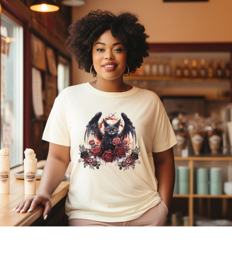 May include: Cream-colored t-shirt featuring a black cat with bat wings, surrounded by red and black roses. The cat has yellow eyes and is set against a red background. The shirt is a casual, everyday wear item.