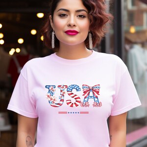 May include: A pink t-shirt with a graphic of the word "USA" made up of red, white, and blue stars and stripes. The letters are decorated with flowers and bows.