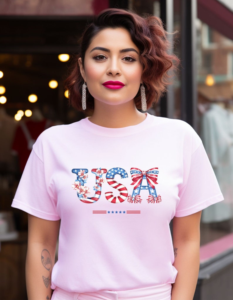 May include: A pink t-shirt with a graphic of the word "USA" made up of red, white, and blue stars and stripes. The letters are decorated with flowers and bows.