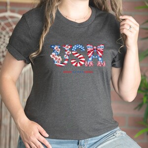 May include: A dark gray t-shirt with a graphic of the word "USA" in red, white, and blue. The letters are decorated with flowers and bows.
