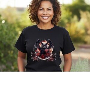 May include: Black t-shirt featuring a graphic of a black cat with bat wings, surrounded by red roses. The cat has yellow eyes and is set against a red and white background. The design is centered on the front of the shirt.