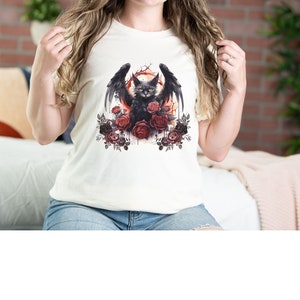 May include: Cream-colored t-shirt featuring a black cat with bat wings, surrounded by red and dark red roses. The cat has yellow eyes and is set against a red and orange background. The shirt is a casual style.