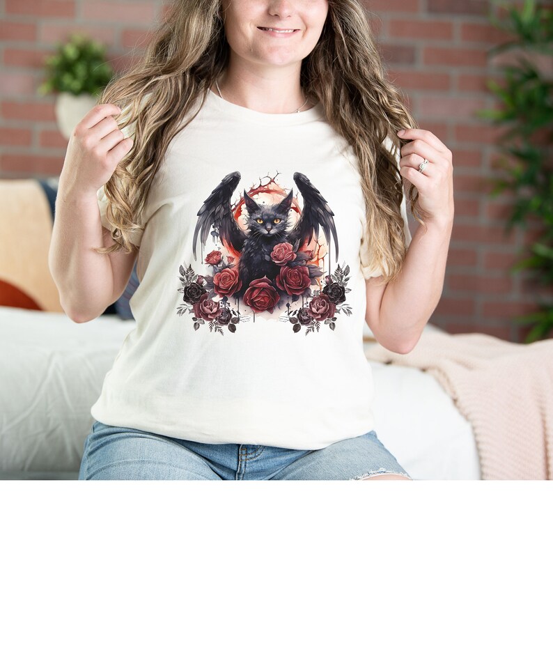 May include: Cream-colored t-shirt featuring a black cat with bat wings, surrounded by red and dark red roses. The cat has yellow eyes and is set against a red and orange background. The shirt is a casual style.