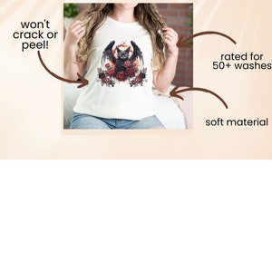 May include: Cream-colored t-shirt featuring a black cat with bat wings, surrounded by red roses, and a crescent moon. The shirt has the text "We use Direct-To-Garment Printing" at the top. The shirt is rated for 50+ washes and made of soft material.