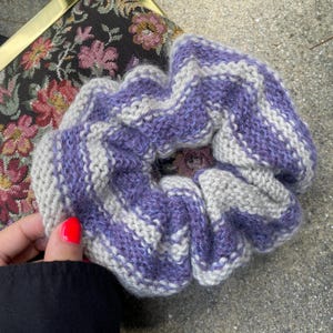May include: A handmade scrunchie in a knitted pattern. The scrunchie features alternating stripes of lavender and light gray. The accessory is designed to hold hair in a ponytail or bun. The scrunchie is made of soft yarn.