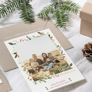 May include: A white Christmas card with a watercolor border of green leaves and red berries. The card features a photo of a family of five smiling in a field. The text "Happy Holidays" is written in gold lettering. The names "John & Mariah, Sam, Hunter & Anna" are written in black lettering below the photo.