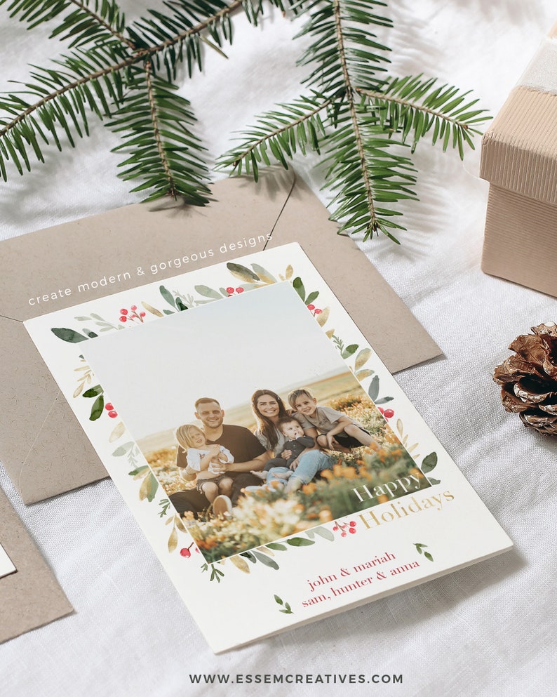 May include: A white Christmas card with a watercolor border of green leaves and red berries. The card features a photo of a family of five smiling in a field. The text "Happy Holidays" is written in gold lettering. The names "John & Mariah, Sam, Hunter & Anna" are written in black lettering below the photo.