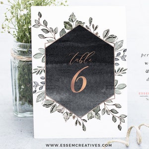 May include: A black and white watercolor table number card with a gold hexagonal frame and greenery. The number 6 is written in gold on the card. The text "table" is written in cursive above the number. The card is perfect for a wedding or event.
