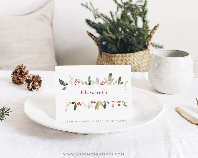 May include: A white place card with a watercolor design of green leaves and red berries. The name "Elizabeth" is written in pink lettering. The text "create clean & festive designs" is printed below the name.