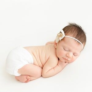 May include: A newborn baby girl sleeping on a white background. She is wearing a white diaper and a white headband with a flower accent.