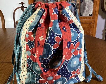 May include: A quilted drawstring bag with a floral pattern in red, blue, and white. The bag has a drawstring closure and a red strap. The bag is made of fabric and has a patchwork design.